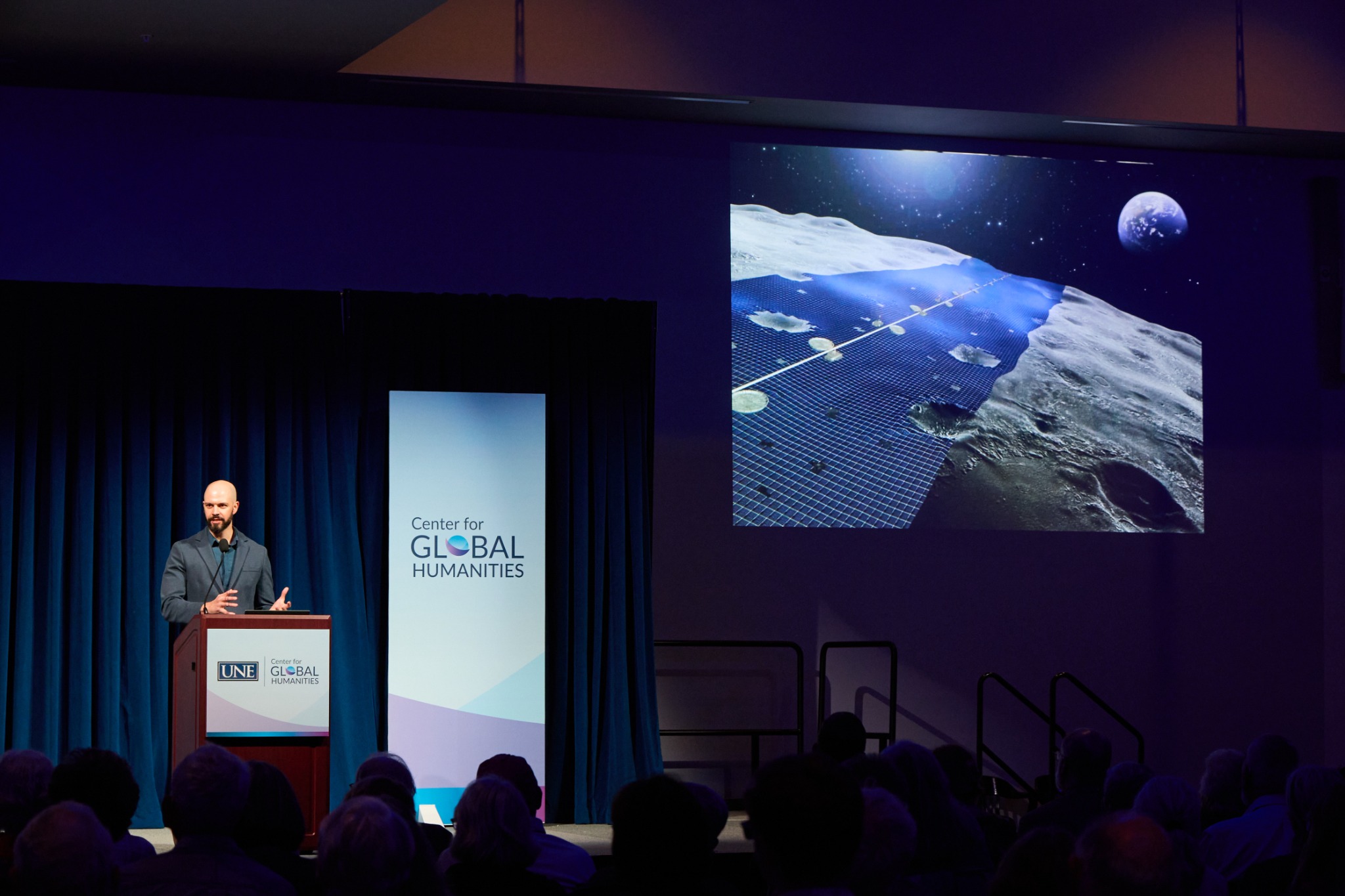 Dagomar Degroot speaking at a podium beside a projected image of a lunar settlement